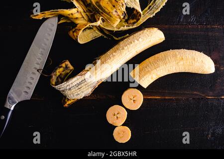 Peeled and Sliced Plantains on a Dark Wood Background: Raw plantains being peeled and sliced with a santoku knife Stock Photo