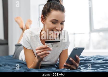 Smiling woman holding credit card and using cellphone while lying on bed at home Stock Photo