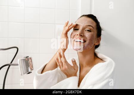 Young smiling young white woman beauty blogger in bahtrobe making a video on mobile phone in the bathroom apply facial scrub Stock Photo