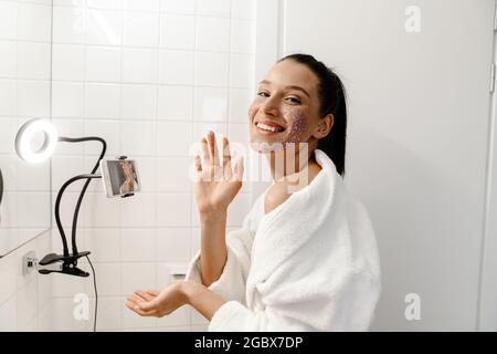 Young smiling young white woman beauty blogger in bahtrobe making a video on mobile phone in the bathroom apply facial scrub Stock Photo