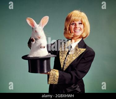 1960s BLONDE FEMALE MAGICIAN LOOKING AT CAMERA SMILING AND LIFTING A WHITE RABBIT OUT OF A TOP HAT - kg3003 HAR001 HARS JOY TRICK SATISFACTION ACTOR FEMALES STUDIO SHOT COPY SPACE HALF-LENGTH LADIES PERSONS CHARACTER PROFESSION ENTERTAINMENT CONFIDENCE EYE CONTACT PERFORMING ARTS OCCUPATION HAPPINESS CHEERFUL PERFORMER AND CAREERS EXCITEMENT PRIDE ENTERTAINER OCCUPATIONS SMILES ACTORS JOYFUL STYLISH SEQUINS ENTERTAINERS PERFORMERS TOP HAT YOUNG ADULT WOMAN CAUCASIAN ETHNICITY HAR001 OLD FASHIONED Stock Photo