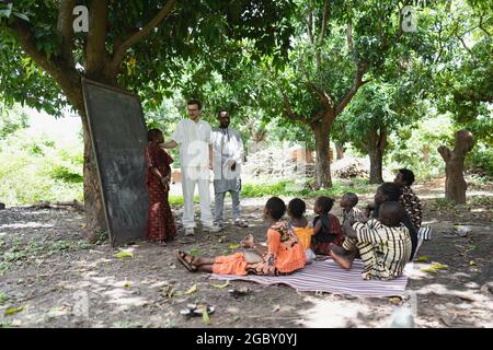 In this image, a tall white volunteer is teaching a group of children in an open air summer camp in a remote village in West Africa Stock Photo