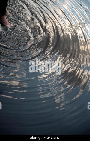 One person's feet in water creating ripples in lake surface with reflected sky Stock Photo