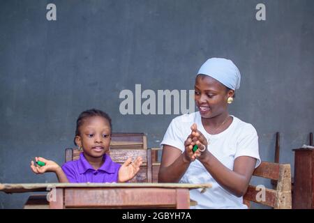 African Nigerian mother or teacher sitting together with her girl child in a classroom, helping her with her studies towards excellence in her, school Stock Photo