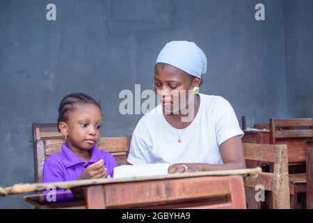 African Nigerian mother or teacher sitting together with her girl child in a classroom, helping her with her studies towards excellence in her, school Stock Photo