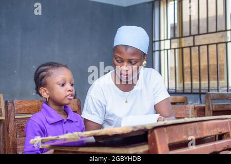 African Nigerian mother or teacher sitting together with her girl child in a classroom, helping her with her studies towards excellence in her, school Stock Photo