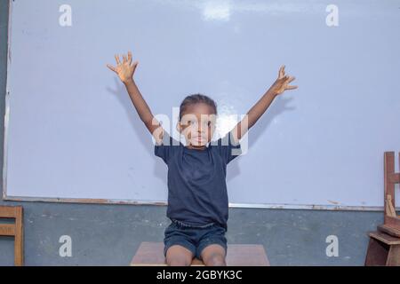 African girl child, pupil or student sitting down in front of a whiteboard and joyfully spreading her hands wide because of excellence in education Stock Photo