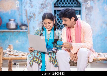 Rural Indian father and young daughter using laptop while sitting on traditional wooden bed outside their house, Happy family looking at computer scre Stock Photo