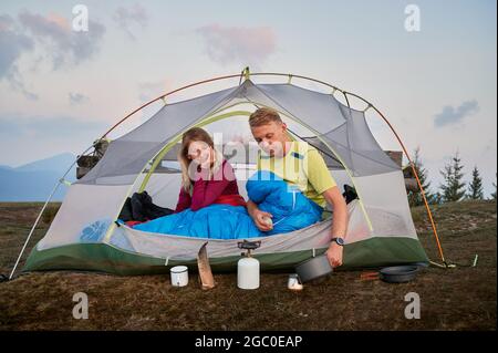Young man in sleeping bag in tent preparing morning coffee for his beloved, waking up after cool night in the mountain fresh air. Tourist breakfast ou Stock Photo