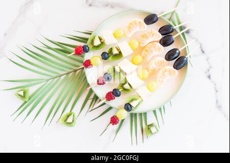 canapes of fresh fruit and berries on a plate. Marble background with palm tree branch. Festive food Stock Photo