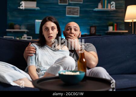 Amazed confused couple watching movie on TV at night and eating popcorn, drinking beer having facial expression. Focused surprised concentrated astonished wife sitting home on confortable couch Stock Photo