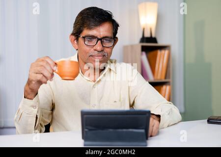 Businessman having coffee while using or reading news, social media and internet on digital tablet at office - concept of taking break during work for Stock Photo