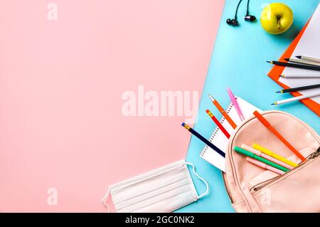 Creative student desk with protective mask, colorful modern school supplies and pink backpack on blue and pink duotone background. Top view. Flat lay. Copy space. Back to school concept Stock Photo