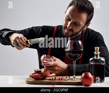 Brutal bearded man in black shirt with striped tie and suspenders sits at table eating fresh pomegranate and juice Stock Photo