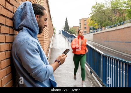 Active man resting after working out outdoors and using his mobile phone. His female friend is still running in the background. Stock Photo