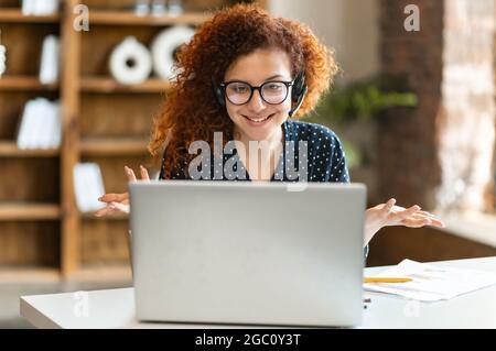 Smiling red-haired curly woman wearing headset looks at the webcam of the laptop and talking online, female employee chatting with colleagues or customer on the distance. Video call, virtual meeting Stock Photo