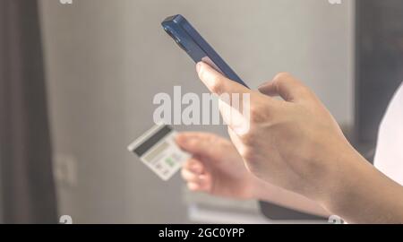 Lady use mobile phone for order online and pay bills with credit card in living room interior at home. Smartphone and credit card in hand close-up, ba Stock Photo