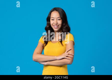Smiling happy mixed race young millennial woman with arms crossed isolated on blue studio background