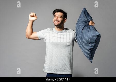 Smiling fresh lively young man holding pillow and stretching after having a good sleep studio shot in isolated light gray background Stock Photo