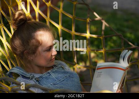 Young blonde woman student smiling Reading A Book While Lying On A Hammock in the garden. Woman relaxing in hammock. Back to school university college Stock Photo