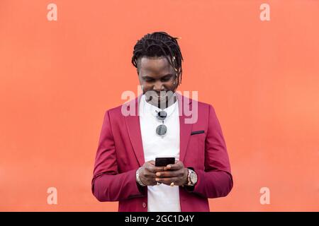 stylish black businessman in suit typing a message on his smart phone in front of a red background, copy space for text, concept of technology and com Stock Photo