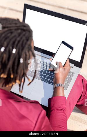 vertical photo of the rear view of an unrecognizable black businessman using a smart phone and computer laptop, technology and remote work concept, co Stock Photo
