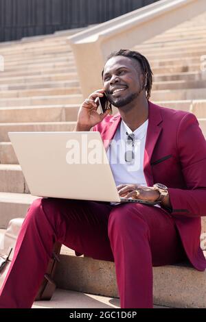 vertical photo of a smiling african businessman working with computer laptop and phone sitting on city stairs, technology and remote work concept Stock Photo
