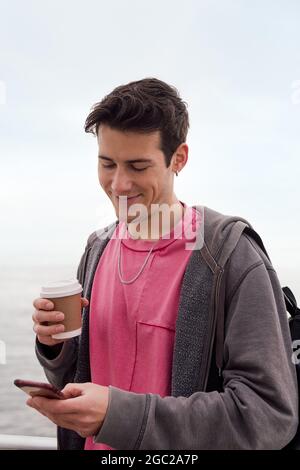 vertical photo of a happy young man with a cup of coffee smiling and using a mobile phone, concept of technology and urban lifestyle Stock Photo