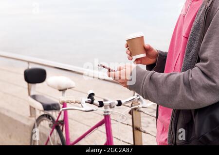 unrecognizable young man with a drink using a mobile phone next to a vintage bike, concept of technology of communication and urban lifestyle, copy sp Stock Photo