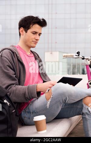 vertical photo of a young student sitting outdoors using tablet next to his bicycle, concept of technology and urban lifestyle, copy space for text Stock Photo