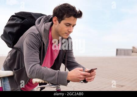 handsome young man with backpack leaning on a bicycle using a mobile phone, concept of communication and ecological urban transportation, copy space f Stock Photo