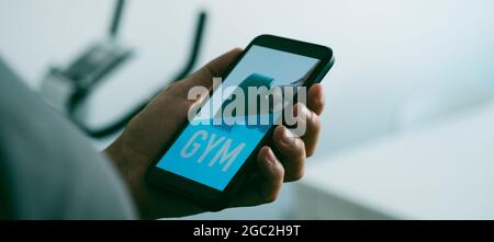 a young caucasian man is looking to his smartphone, that reads gym in its screen, next to a stationary bicycle, in a panoramic format to use as web ba Stock Photo