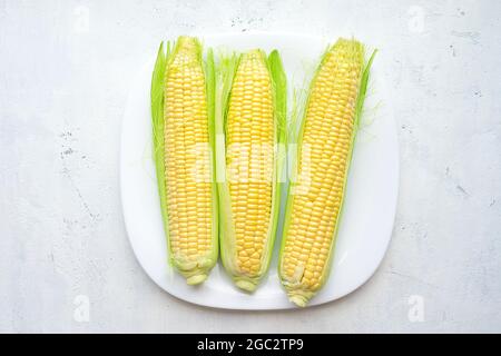 Fresh corn ears with green leaves on a white square plate on a light vintage background. Top view Stock Photo