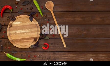 Handmade round wooden plate and spoon with spices, pepper, basil leaves and anise on brown wooden background. Top view, flat lay, copy space Stock Photo