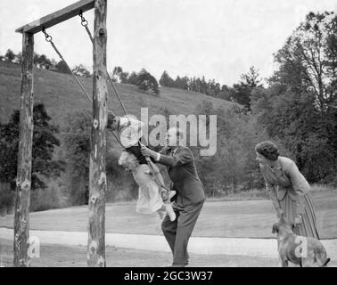 The Royal Family at Balmoral, Scotland - June 1956 Prince Philip, The Duke of Edinburgh pushes his children, Princess Anne and Prince Charles, on a swing, with Queen Elizabeth looking leaning down to a pet dog. Stock Photo