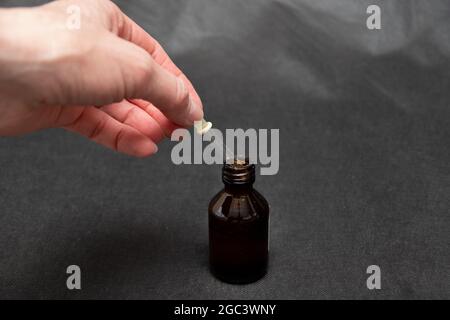 A hand holds a glass dropper with a yellow rubber tip with liquid over a dark brown bottle on a dark trendy background close-up.