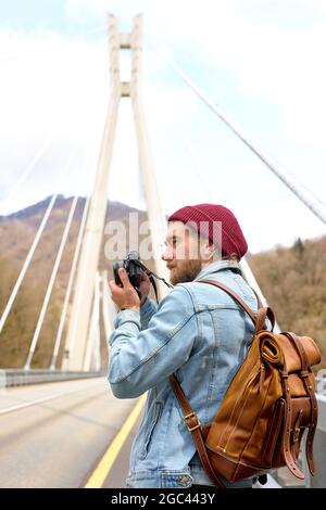 Confident photographer hipster caucasian guy walking on bridge in urban area taking photo on camera, wearing denim jacket, looking at side, alone outd Stock Photo