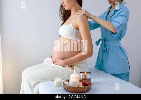 Masseur physiotherapist in blue uniform massaging back of pregnant woman in spa center, side view. Mom-to-be preparing to give birth in a short time. Stock Photo