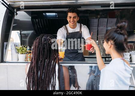 Back view of two females buying drinks from a male owner at a food truck. Smiling entrepreneur giving drinks to clients. Stock Photo
