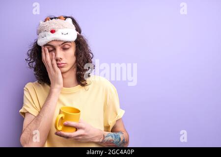 Brunette curly hair caucasian man has no enough sleep, want to go in bed in the morning, dressed casually with eyemask for sleeping, holding yellow mu Stock Photo