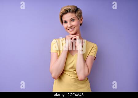 Pleasant Cute Female With Short Hair Posing At Camera Isolated On Purple Background, Looking Kind And Pleasant, Keeping Hands Together, Portrait of Ca Stock Photo