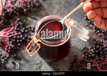 Hand holding a spoon with homemade black elder syrup, with fresh elderberries in the background Stock Photo