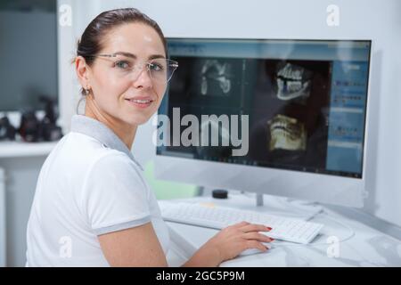Charming female dentist smiling joyfully while working with x-ray scans on her computer Stock Photo