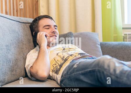 Young Caucasian man lie relax on sofa at home use modern smartphone gadget, relaxed millennial male rest on comfortable couch in living room, technolo Stock Photo