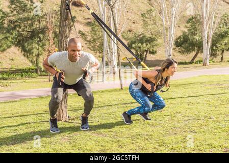 Athletic partners doing squatting back exercise with fitness straps in park Stock Photo