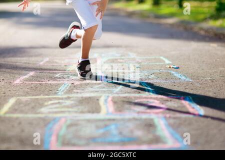 Kids play hopscotch in summer park. Healthy active outdoor game. Children playing and drawing with chalk on suburban city street. Boy and girl jump. Stock Photo