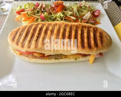 A Ham and cheese panini sandwich served with fresh vegetable salad on the side served on a white plate. Stock Photo