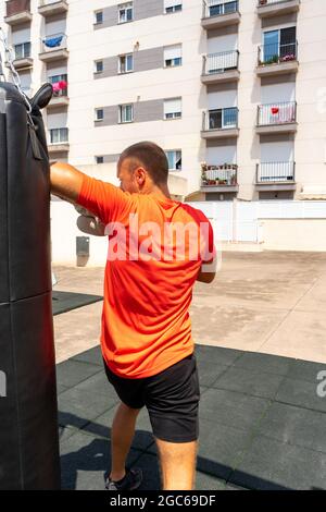 Street Fighter in Black Clothes and Bandages on the Wrist Boxing in Punching Bag Outdoors. Young Man Doing Box Training and Practicing His Punches at Stock Photo