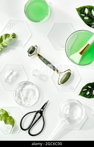 Self made moisturizer, green jade face roller and scissors with pieces of ice. Exotic monstera leaves and water drops on white background. Facial Stock Photo