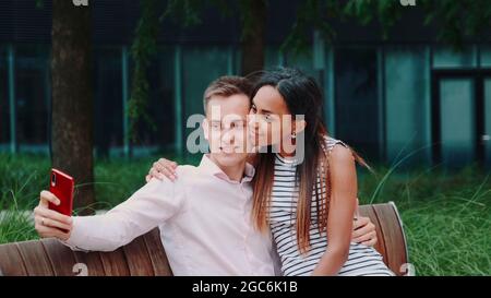 Romantic multiethnic couple sitting on bench in park and taking selfie. They smiling to the camera. Stock Photo